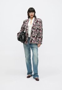 Σακάκι με ανθισμένο μοτίβο σε σκούρες αποχρώσεις, συνδυασμένο με λευκό πουκάμισο, ανοιχτόχρωμα τζιν και σκουρόχρωμα παπούτσια. Το μοντέλο κρατά μια μαύρη τσάντα.