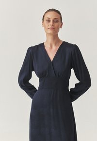 Vestido azul marino con mangas largas abullonadas y un escote en V profundo. Presenta una cintura ajustada y una textura suave con un patrón sutil.