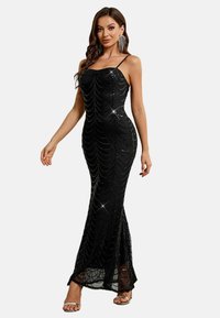 Eine Frau trägt ein ärmelloses, schwarzes, mit Pailletten besetztes Abendkleid mit dünnen Trägern und silbernen High Heels und posiert vor einem weißen Hintergrund.