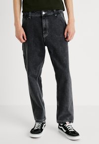 Persona vestida con pantalones vaqueros negros de corte holgado y zapatillas bajas en blanco y negro, de pie frente a un fondo liso.