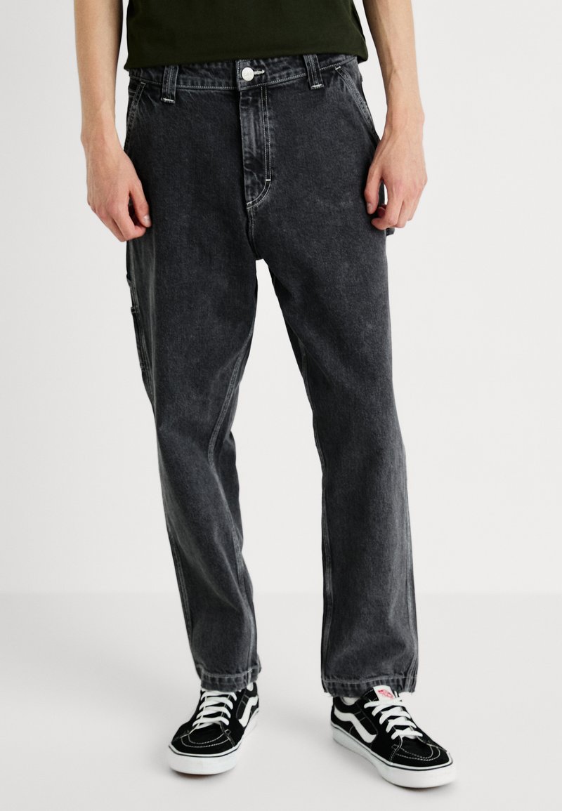 Persona vestida con pantalones vaqueros negros de corte holgado y zapatillas bajas en blanco y negro, de pie frente a un fondo liso.