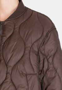 Primo piano di una giacca marrone trapuntata con cuciture a motivo ondulato e chiusura frontale con cerniera, indossata sulla parte superiore del busto e sulla spalla di una persona.