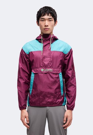 Mladý muž oblečený do fialovo-tyrkysovej bundy Columbia Sportswear proti vetru a sivých nohavíc stojí na jednoduchom svetlom pozadí.