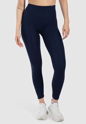 Vrouw in marineblauwe high-waist leggings en witte sneakers, staat met ontspannen armen langs haar lichaam tegen een neutrale achtergrond.