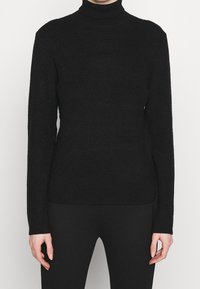 Femme portant un pull à col roulé en tricot côtelé noir et un pantalon noir ajusté, debout devant un fond clair uni.