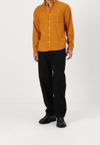 Camicia button-up a scacchi arancione con maniche lunghe, dotata di taschino sul petto, abbinata a pantaloni neri e scarpe nere.