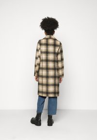 Cappotto a quadri in beige, nero e marrone, con silhouette dritta, rever a punta e maniche lunghe, abbinato a jeans blu cropped e stivali neri.