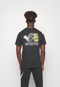 T-shirt en coton noir avec un graphique de limace de dessin animé et de pneu. Le texte indique "NIKE BODY SHOP", avec des accents jaunes et un motif à damier en dessous.