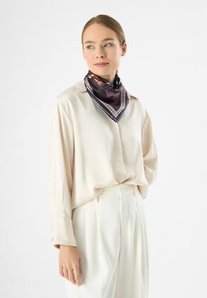 Crème-kleurige blouse met lange mouwen en een relaxte pasvorm, gecombineerd met hoog-getailleerde witte broeken. Een gemusterde zijden sjaal is om de hals geknoopt.