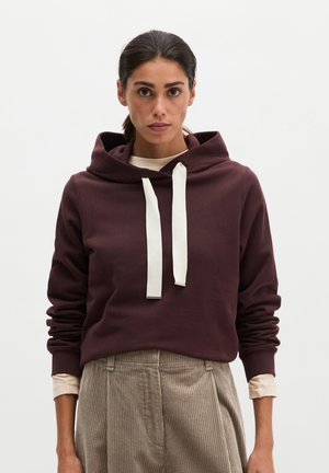 Frau mit dunklem, zurückgebundenem Haar, die einen dunkelroten Hoodie mit weißen Kordeln und beige Cordhosen vor einem schlichten weißen Hintergrund trägt.