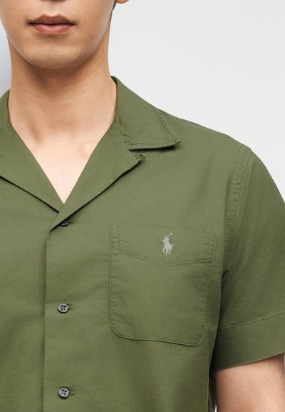Άνδρας που φοράει πουκάμισο με κοντά μανίκια σε ελιά πράσινο χρώμα, με τσέπη στο στήθος που φέρει ένα μικρό κεντητό λογότυπο παίκτη πόλο.