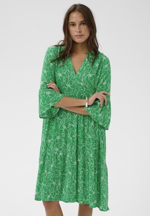 Žena s dlhými hnedými vlasmi, oblečená v zelenej kvetinovej šatách do kolien s tromi štvrťovými rukávmi, stojaca so založenými rukami na jednoduchom pozadí.