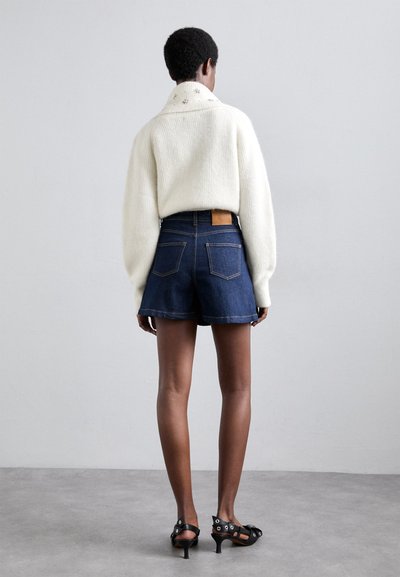 White, textured sweater with a wide collar, paired with dark denim shorts featuring a back patch and two pockets. Black heeled sandals.