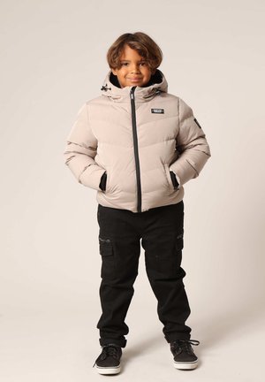 Lichtbeige pufferjack met capuchon, ritssluiting, zwarte accenten en elastische manchetten, gecombineerd met zwarte cargobroek en zwarte sneakers.