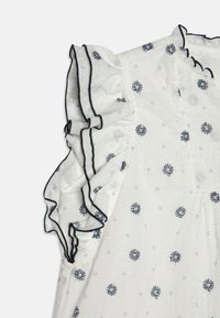 Blouse blanche avec des manches noires à volants, ornée d'une broderie florale bleue, en tissu texturé avec de petits motifs en pointillés, et un contraste avec un bord foncé.
