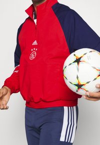 Chaqueta roja con cremallera y mangas azul marino, con un logo y tres estrellas. Pantalones azules con rayas blancas, sosteniendo un balón de fútbol con un estampado.