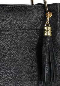 Zwart ledertas met textuur, voorzien van een gouden metalen accent en een zwarte kwast. Het ontwerp kenmerkt zich door minimalistische lijnen en een licht gestructureerde vorm.