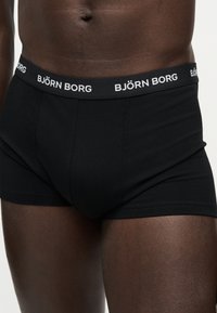 Φωτογραφία ανδρικού κορμού με σκούρο δέρμα που φοράει μαύρο μπόξερ Björn Borg με λογότυπο στη ζώνη, σε ουδέτερο φόντο.