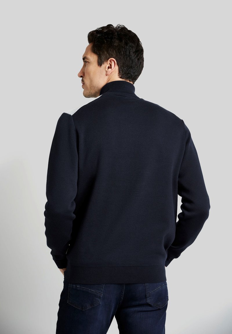 Marineblauer Pullover mit hohem Kragen, gerippten Bündchen und Saum, der überarbeitete Schulterakzente aufweist. Hergestellt aus weichem Strickmaterial.