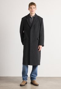 Langer schwarzer Wollmisch-Mantel mit Reverskragen, Fronttaschen und strukturiertem Finish, kombiniert mit einem grauen Hemd und blauen Jeans.