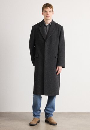 Abrigo largo negro de mezcla de lana con cuello con muesca, bolsillos frontales y un acabado texturizado, combinado con una camisa gris y vaqueros azules.