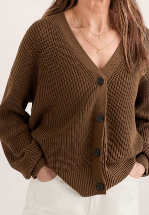 Cardigan en maille épaisse marron avec des boutons noirs, porté avec un pantalon blanc, assorti à des colliers dorés superposés ornés d'oiseaux.