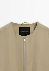 Veste beige à col rond avec une fermeture éclair argentée visible, dotée d'une étiquette noire "Massimo Dutti" à l'intérieur du col et d'une boucle en tissu pour la suspendre.