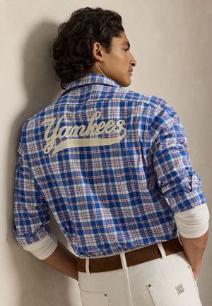 Muž stoji bokom, má na sebe modrú kockovanú košeľu s textom „Yankees“ na chrbte, skombinovanú s bielymi nohavicami a hnedým opaskom.