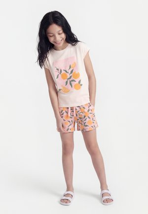 Ragazza giovane che sorride, indossa una maglietta rosa chiaro e pantaloncini con motivi di frutta arancione e foglie, in piedi con le mani vicino alle tasche e sandali bianchi.