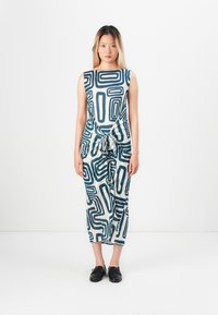 Kobieta stoi na białym tle, ubrana w bezrękawnikową sukienkę w niebieskie abstrakcyjne wzory geometryczne i czarne buty wsuwane.