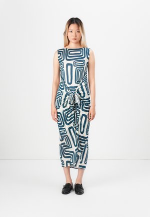 Kvinde står foran hvid baggrund iført en ærmeløs kjole med blå abstrakte geometriske mønstre og sorte slip-on sko.