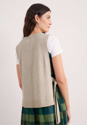 Maglione senza maniche di un beige chiaro in morbido knit con orlo a coste, abbinato a un vestito a quadri verde e blu. Presenta una cintura in vita.