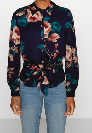 Marineblå blomsterprint skjorte med knapper, lange ærmer og bundet talje, kombineret med lyseblå denimjeans, forfra set fra hals til hofter.