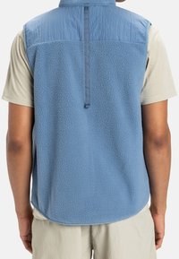 Fleecová vesta světle modré barvy, s texturovaným zadním dílem, hladkým horním panelem a předním zipem. Lem má mírně zakřivený tvar a je nezačištěný.