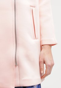 Abrigo rosa claro hecho de un tejido suave que cuenta con una cremallera central, bolsillos laterales y mangas de tres cuartos, con una textura suave y un diseño moderno.