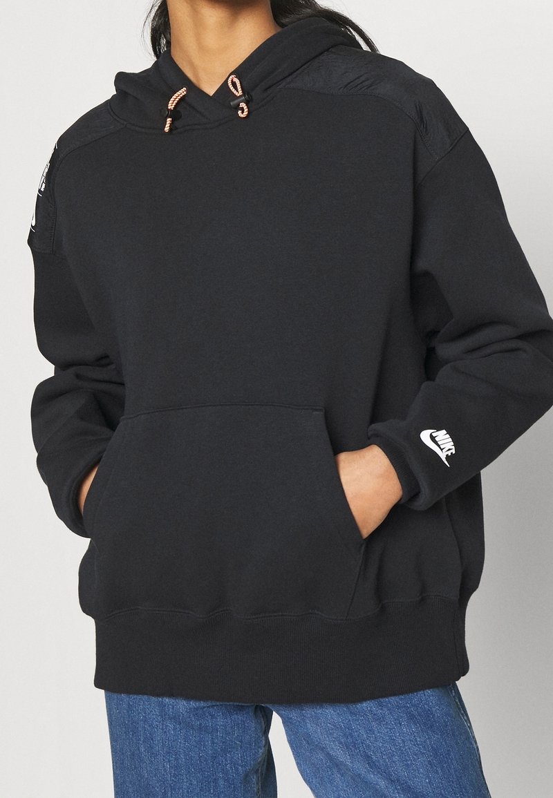 Svart hoodie i mjukt material, med en framficka, dragsko i huven med metallspetsar, och en vit Nike-logotyp på ärmen.