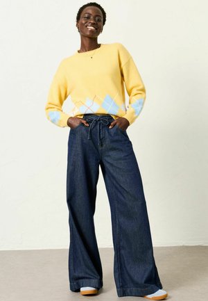 Lächende Person mit einem gelben Pullover mit blauem Rautenmuster und weiten Jeanshosen posiert mit den Händen in den Taschen.