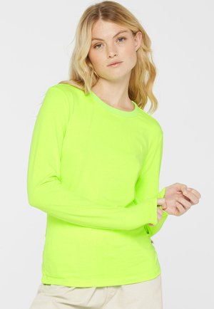 Frau mit blondem Haar, die ein leuchtend neongelbes Langarmshirt und hellfarbene Hosen vor einem schlichten Hintergrund trägt.