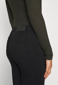 Jeans de mezclilla negra con una textura suave, de cintura alta y bolsillos traseros. Una etiqueta de cuero oscuro es visible en la cintura. Mangas largas en color verde oliva.