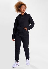 Svart huvtröja med framficka och snörning, tillsammans med svarta sweatpants med en logotyp, kompletterat med vita sneakers.