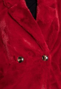 Lähikuva punaisesta kaksirivisestä tekoturkistakista, jossa on ruskeat pyöreät napit ja leveät kaulukset, mustan topin päällä.