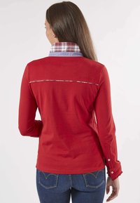 Chemise rouge à manches longues avec un col à carreaux, dotée d'une broderie de marque dans le dos et de détails en boutons sur les poignets. Texture lisse.