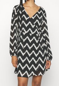 Vit och svart chevronmönstrad omslagklänning med långa ärmar, v-ringning och en knytning i midjan, tillverkad av lättviktsmaterial.