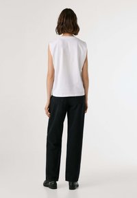Donna con i capelli corti in piedi, vestita con una maglietta bianca senza maniche, pantaloni neri a gamba larga e scarpe nere, rivolta verso uno sfondo chiaro e semplice.