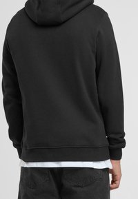 Felpa nera con cappuccio in tessuto morbido, dotata di coulisse, polsini e orlo a coste. Indossata sopra una camicia bianca, con un design minimalista.