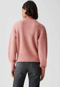 Person, die einen gerippten, staubigen rosa Pullover und schwarze Jeans trägt, von hinten gegen einen schlichten hellen Hintergrund gezeigt.