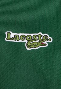 Polo shirt verde in cotone con logo "Lacoste" ricamato in bianco ed emblema del coccodrillo. Texture a coste e design aderente.