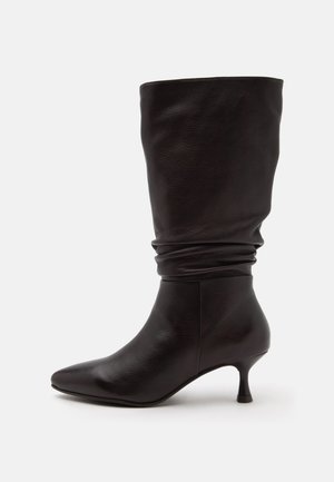Bota de cuero negra hasta la mitad de la pantorrilla con caña caída, puntera puntiaguda y tacón bajo tipo kitten sobre un fondo claro y liso.