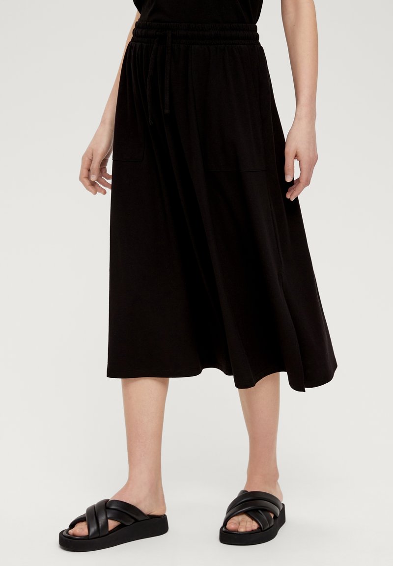 Personne portant une jupe midi noire à poches, des sandales noires à brides croisées et un haut noir, debout devant un fond clair et uni.