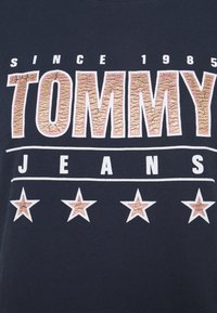 Námořnické tričko s texturovaným zlatým potiskem "TOMMY", bílými hvězdami pod ním a "JEANS" v bílé barvě, s kulatým výstřihem a krátkými rukávy.